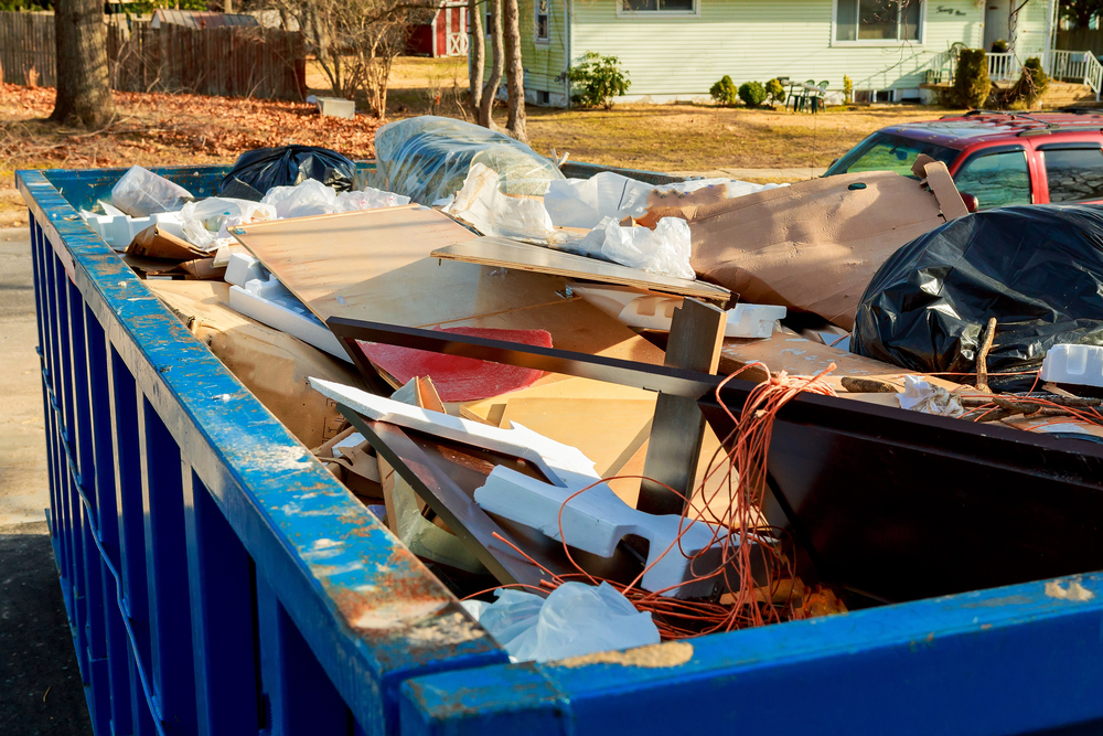 A blue dumpster filled with various discarded materials, including wood, plastic, wire, and trash bags, is situated outdoors near houses and parked cars.