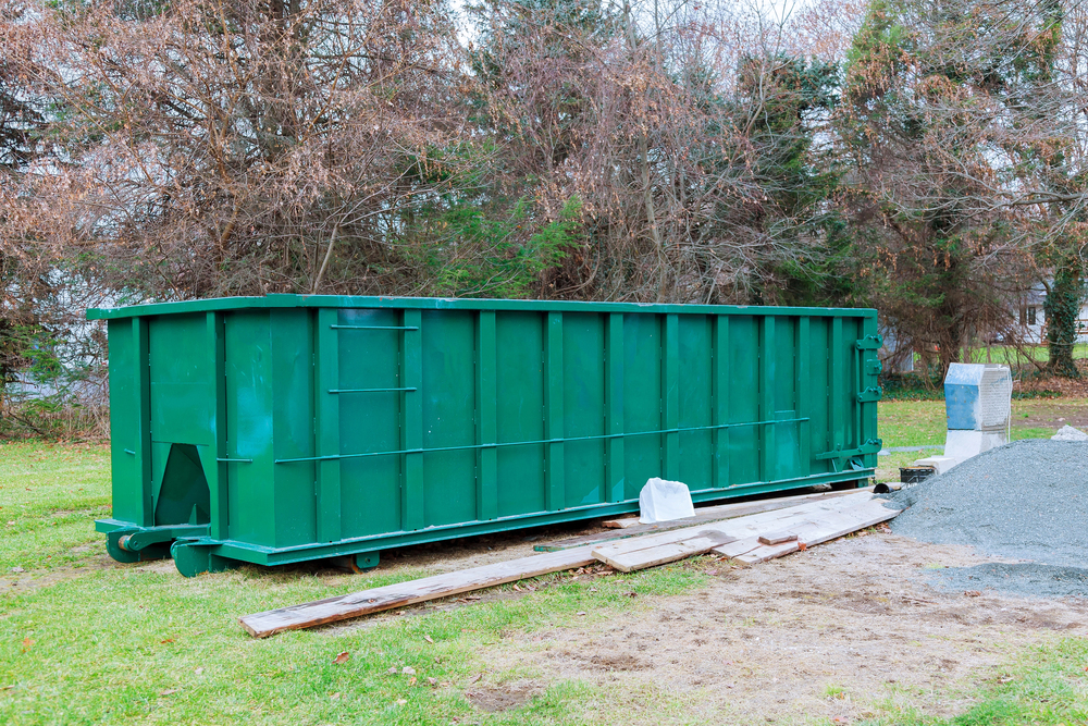 A large green dumpster sits on grass beside wooden planks, a concrete block, and a gravel pile, with leafless trees in the background.