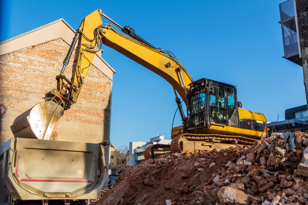 A yellow excavator loads debris into a dump truck at a construction or demolition site on a clear day.