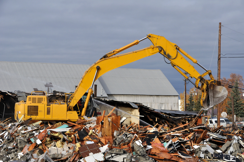 A yellow excavator is clearing debris from a demolished building, with piles of rubble and metal in the foreground and industrial buildings in the background.