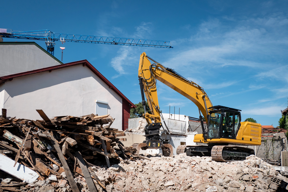 A yellow excavator is clearing debris from a demolished building site, with a crane and intact buildings visible in the background under a blue sky.