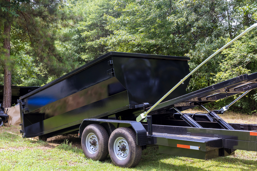 A black dump trailer with dual axles is tilted to unload contents, parked on grass near trees.