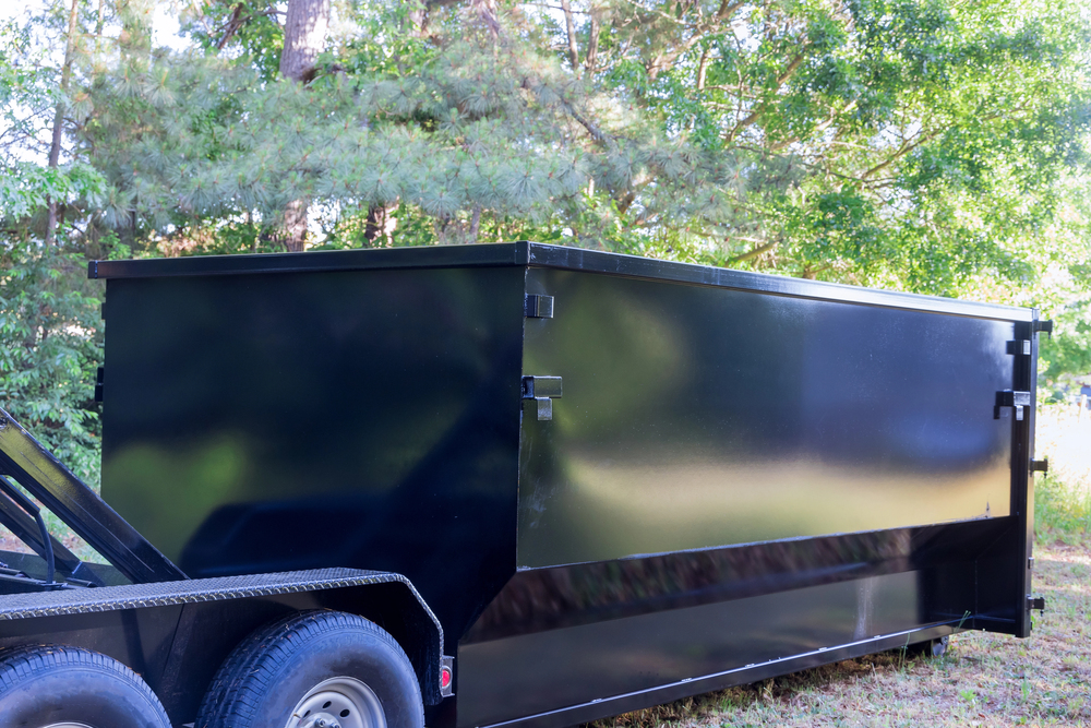 A large, black, rectangular metal dumpster sits outdoors on a trailer, surrounded by trees and greenery in the background.