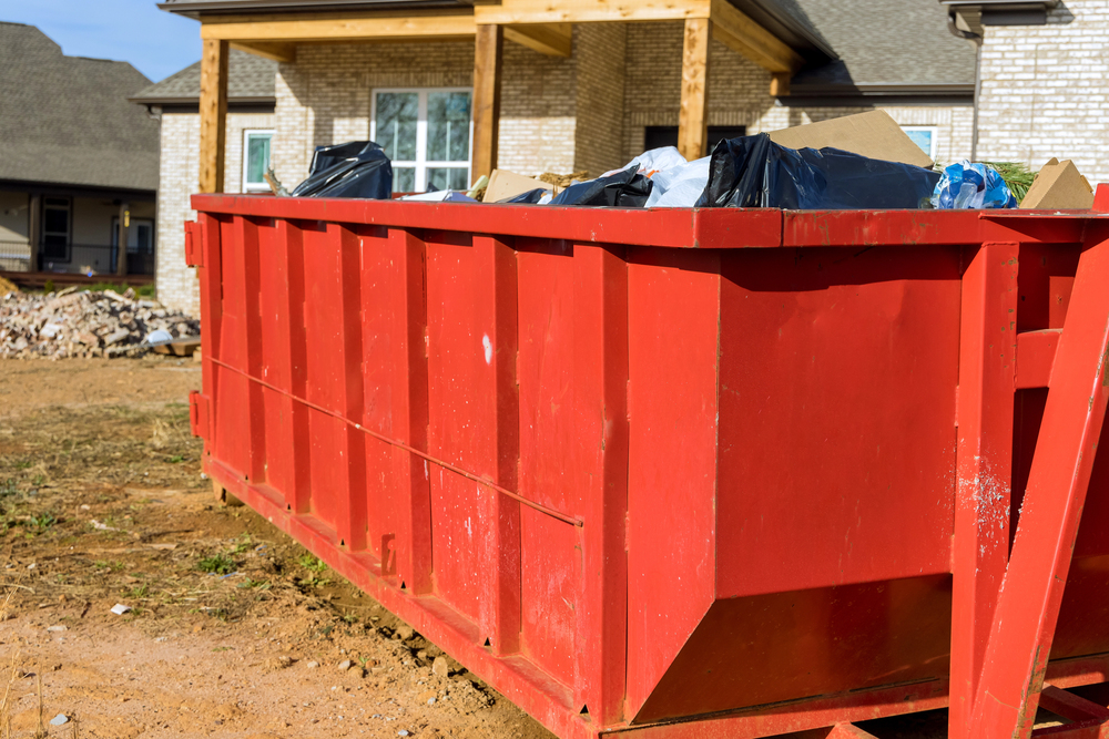 A large red dumpster filled with bags of trash and debris sits outside a brick house under construction or renovation.