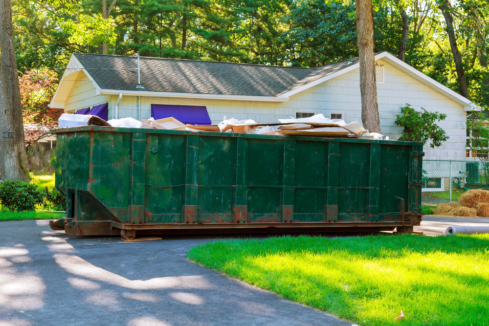 Large green dumpster filled with debris sits on a driveway in front of a single-story house surrounded by trees and grass.
