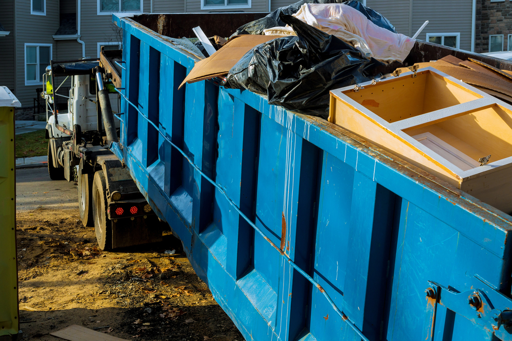 A large blue dumpster filled with construction debris, including wood, plastic bags, and a cabinet, sits beside a truck on a dirt surface near residential buildings.
