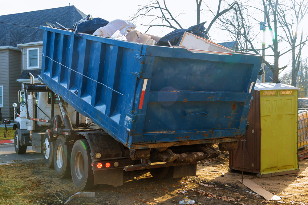A large blue dumpster filled with debris is being loaded onto a truck at a residential construction site, next to a yellow portable toilet.