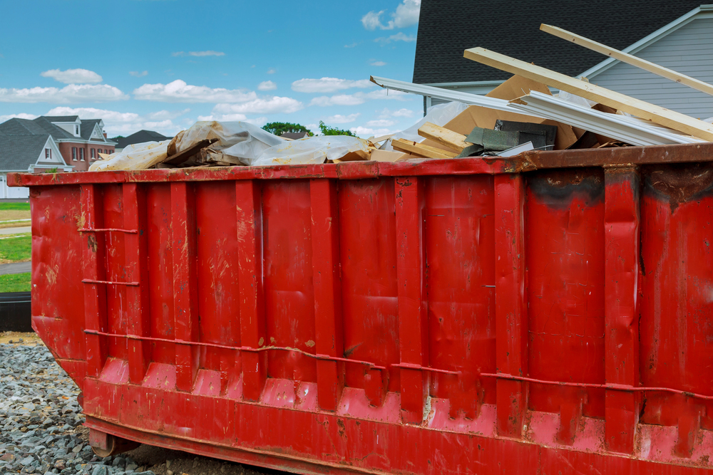 A large red dumpster filled with construction debris and wood sits beside a house under a partly cloudy sky.